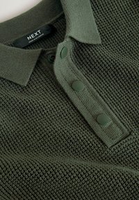 Polo shirt verde a maglia con un motivo testurizzato, colletto e tre bottoni verde scuro. L'etichetta di cura mostra 'NEXT, made in Myanmar.'
