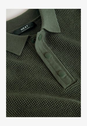 Polo verde de punto con un patrón texturizado, un cuello y tres botones verde oscuro. La etiqueta de cuidado muestra 'NEXT, fabricado en Myanmar.'