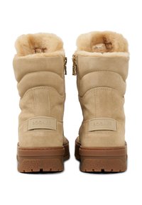 Béžové semišové boty s plyšovou kožešinovou výstelkou, dvojitými bočními zipy, texturovanými podrážkami a gumovou střední podrážkou. Na zadní straně je logo.