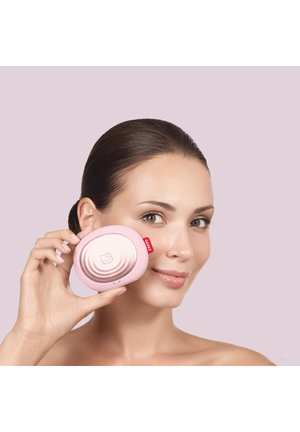 Kvinde med trukket hår tilbage holder et lyserødt kompakt spejl med et Hello Kitty-design tæt på sin kind mod en lys lilla baggrund.