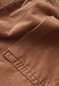 Tissu en velours côtelé marron avec une texture côtelée visible. Présente un bord replié et un détail de couture. Aspect doux et souple.