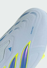 Gros plan sur la tige d'une chaussure de sport blanche avec une surface texturée, le texte bleu "Nanostrike" et des accents de design irisés en violet et jaune.