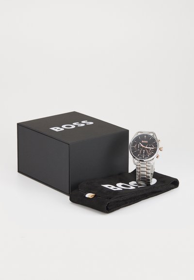 Montre en acier inoxydable argenté avec un cadran noir, accents en or rose, présentée à côté d'une boîte noire et d'une pochette en suédine protectrice avec logo.