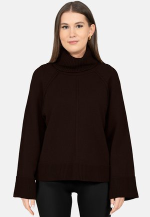 Junge Frau trägt einen dunkelbraunen Rollkragenpullover mit breiten Bündchen und schwarze Hosen, steht vor einem einfarbigen hellen Hintergrund.