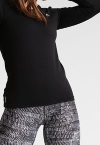 Schwarzes langärmliges Sportoberteil kombiniert mit engen Leggings, die ein monochromes geometrisches Muster aufweisen. Beide Kleidungsstücke bestehen aus dehnbarem Material.