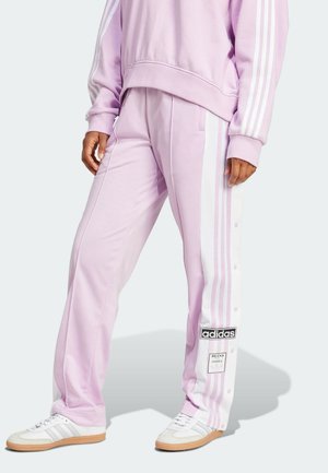 Osoba ubrana w jasnofioletowe spodnie dresowe Adidas z białymi paskami po bokach oraz białe buty sportowe Adidas z gumowymi podeszwami.