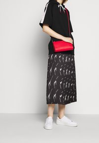 Kvinde iført en sort hættetrøje med korte ærmer, sort plisseret nederdel med hvidt kradsemønster, hvide sneakers og holder en rød crossbody taske.