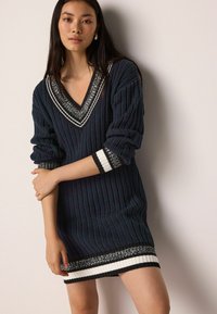 Rochie tricotată cu decolteu adânc în V, de culoare bleumarin închis, textură ribatată și manșete și tiv decorative cu dungi, având accente albe și negre.