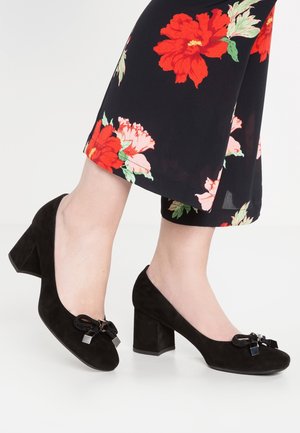 Pieds chaussés de chaussures à talons blocs en daim noir avec des nœuds décoratifs, assorties à un pantalon noir raccourci orné de grandes impressions florales rouges et roses.