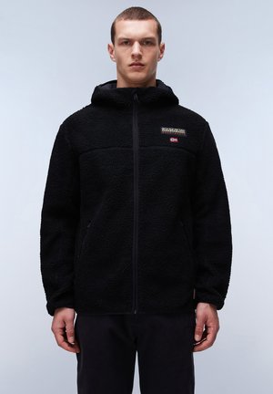 Młody mężczyzna noszący czarną polarową kurtkę z kapturem i zamkiem z przodu, z małą naszywką z logo oraz norweską flagą na piersi.