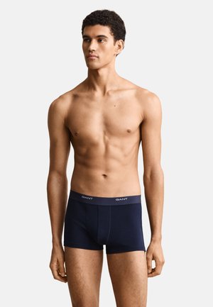Young man with short curly hair wearing navy blue boxer briefs with a branded waistband, standing against a plain white background.