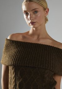 Brązowy sweter typu off-the-shoulder z prążkowanym wykończeniem przy dekolcie i wzorem w warkocze na korpusie, prezentujący ciepły, ziemisty odcień.