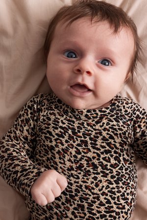 Bébé aux yeux bleus portant un body à manches longues à imprimé léopard, allongé sur une couverture beige, regardant vers le haut avec la bouche légèrement ouverte.