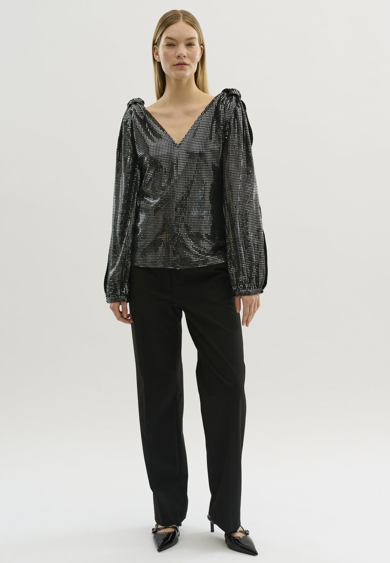 Blusa nera con paillettes, scollo a V e maniche a sbuffo, abbinata a pantaloni neri a gamba larga e décolleté neri a punta. Texture liscia, superficie riflettente.