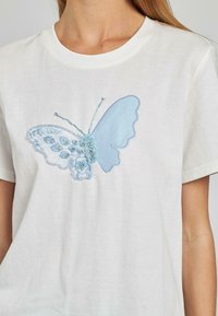 T-shirt in cotone bianco con un motivo ricamato di una farfalla azzurro chiaro, arricchito da dettagli in perline e cuciture dettagliate sulle ali.