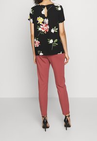 Chemisier noir à motif floral avec manches courtes, présentant une ouverture en forme de clé dans le dos ; associé à un pantalon rose ajusté et des chaussures à talons noires.