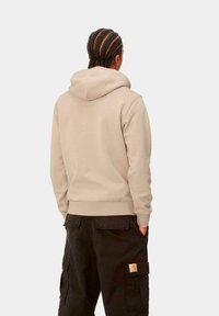 Sudadera beige con capucha ajustable, puños y dobladillo acanalados. Combinada con pantalones cargo negros que tienen bolsillos laterales y un parche con logo.