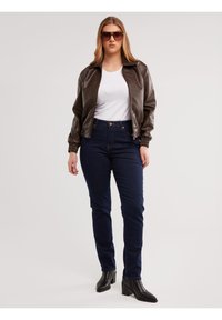 Sieviete, tērpusies brūnā ādas jakā, baltā kreklā, tumši zilās džinsās, melnās zābakos ar papēdi un lielās brūnās saulesbrillēs, stāvot pret balto fonu.