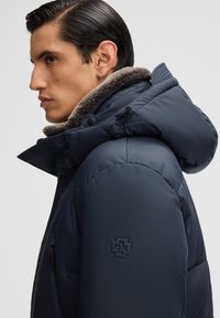 Marineblauwe pufferjack met een met bont gevoerde capuchon, voorzien van een ritszak en een getextureerd logo-accent op de mouw. Glad, gewatteerd materiaal.