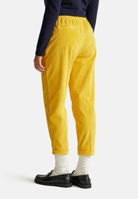 Geel corduroy broek met een relaxte pasvorm, voorzien van een elastische tailleband en opgerolde boorden. Gemaakt met witte sokken en zwarte instappers.