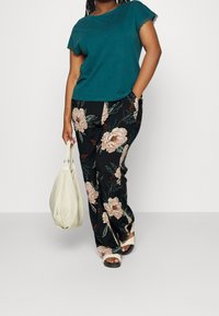 Haut teal à manches courtes avec des dentelles, associé à un pantalon floral noir avec de grandes fleurs beiges. Tient un sac à main crème ; porte des sandales plates.