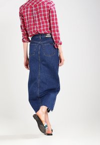 Falda maxi de mezclilla con dobladillo sin acabar, con costuras laterales y dos bolsillos traseros, combinada con una camisa de cuadros roja y blanca. Sandalias planas negras.