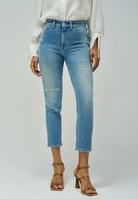 Jeans de tiro alto en azul claro con un diseño ajustado, detalles desgastados en las rodillas y tobillos entallados. La tela es de mezclilla con una textura suave.