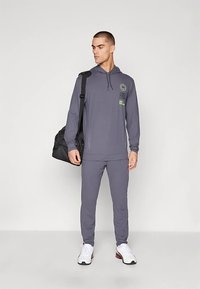 Hombre con sudadera con capucha gris y pantalones a juego, sosteniendo una bolsa de deporte negra, vistiendo zapatillas deportivas blancas, de pie contra un fondo blanco.