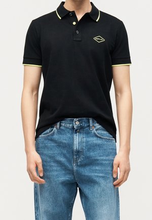 Mand iført sort polo shirt med gul kant og Replay-logo kombineret med blå denimjeans mod ensfarvet baggrund.