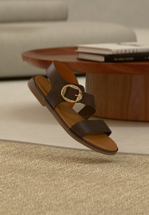 Sandalia de cuero marrón con correas anchas y una gran hebilla de oro, flotando sobre una alfombra beige texturizada cerca de una mesa de centro de madera.