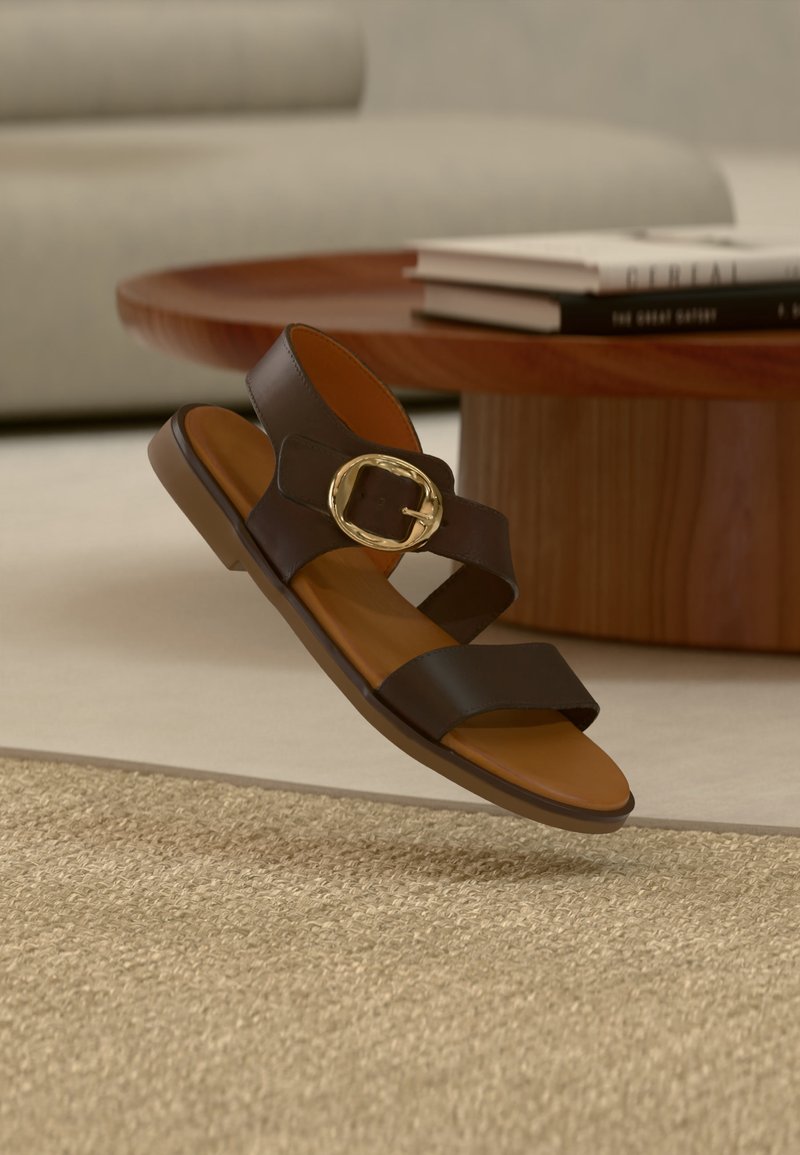 Sandale en cuir marron avec des larges brides et une grande boucle dorée, flottant au-dessus d'un tapis beige texturé près d'une table basse en bois.