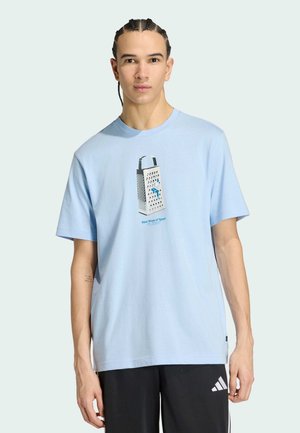 Giovane uomo con i capelli intrecciati che indossa una maglietta azzurro chiaro con una grafica di una grattugia per formaggio e pantaloni neri Adidas.