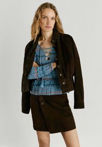 Blonde vrouw met golvend haar draagt een blauw gebreid topje met veters, een donker suède jasje met knopen over haar schouders en een bijpassende donkere suède rok met knopen.