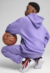Lilla hættetrøje og sweatpants i blødt stof. Hvide og røde atletiske sko med sorte detaljer. Basketball med sort "PUMA" logo.