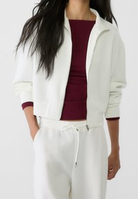 Chaqueta blanca corta con cremallera y un cuello alto, combinada con una blusa ajustada de un burdeos profundo. Pantalones de tela suave y cintura elástica en blanco.