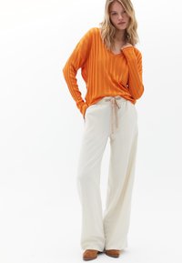 Maglietta a maniche lunghe arancione a coste abbinata a pantaloni larghi color crema con vita regolabile. Scarpe marroni completano l'outfit.