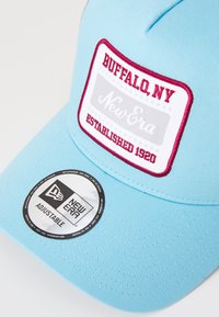 Gorra ajustable de color azul claro con un parche rojo y blanco que presenta el texto "BUFFALO, NY" y "New Era Established 1920." Etiqueta circular de la marca en el ala.