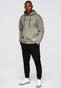 Mies, joka käyttää harmaata hupparia, jossa on teksti "ombre wear", mustia cargo-housuja ja valkoisia lenkkareita, seisoo kädet taskuissa tasaisella taustalla.