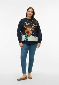 Granatowy sweter z wzorem renifera z czerwonym nosem, płatkami śniegu i zielonym, wzorzystym szalikiem; w połączeniu z niebieskimi dżinsami i brązowymi balerinami.