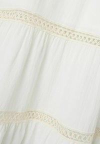 Hvid, tekstureret stof med et lagdelt design, der har en dekorativ, lys beige cirkulær blondertrim langs kanten. Blødt og flydende udseende.