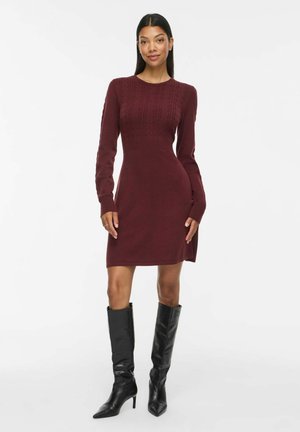 Donna che indossa un vestito aderente in maglia bordeaux a maniche lunghe con motivo a trecce e stivali neri alti al ginocchio con tacco, in piedi su sfondo bianco.