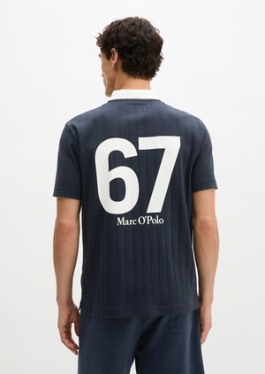 Homme portant une chemise bleu marine à manches courtes avec col blanc, grand numéro blanc 67 et texte « Marc O'Polo » dans le dos.