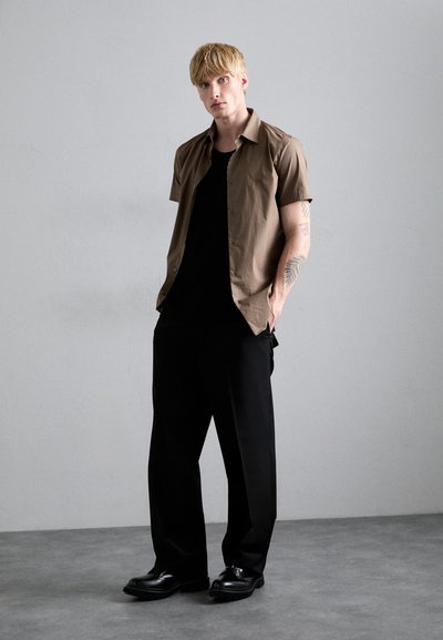 Bruine korte mouwen shirt, zwarte t-shirt eronder, zwarte wijde broek, en zwarte schoenen. Achtergrond is grijs, met een gladde textuur.