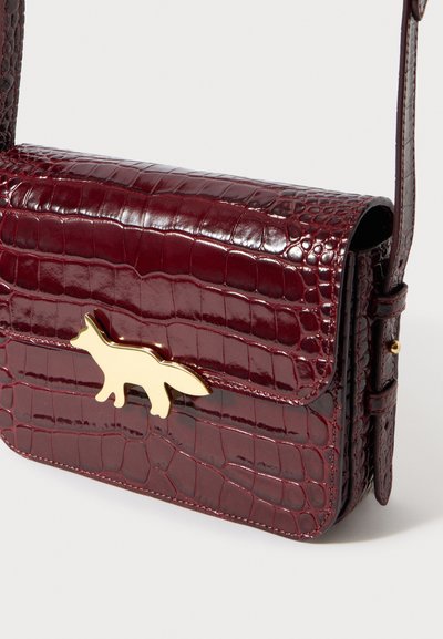 Τσαντάκι χιαστί από δέρμα μπορντό με ανάγλυφο σχέδιο κροκόδειλου, με χρυσό κούμπωμα σε σχήμα αλεπούς και ρυθμιζόμενο λουράκι, πάνω σε λευκό φόντο.