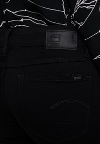 Los vaqueros de mezclilla negra cuentan con una etiqueta de cuero con la marca, costuras en contraste y un detalle en el bolsillo trasero. La textura es suave y duradera.