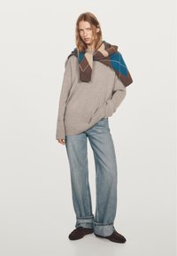 Maglione grigio oversize con cappuccio, abbinato a jeans larghi blu chiaro. Una sciarpa a motivo argyle multicolore pende sulle spalle.