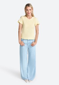 Top a righe giallo chiaro con maniche a balze corte, abbinato a jeans a gamba larga blu chiaro. Modella in piedi con le mani in tasca.