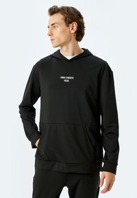 Sudadera con capucha negra de tejido suave, que cuenta con un bolsillo canguro en la parte frontal y texto blanco que dice "FUERZA ENFOQUE FOCO" en el pecho.