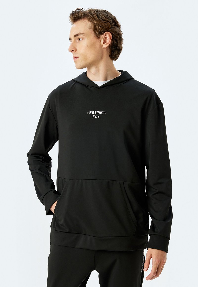 Sudadera con capucha negra de tejido suave, que cuenta con un bolsillo canguro en la parte frontal y texto blanco que dice "FUERZA ENFOQUE FOCO" en el pecho.