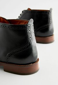Botas de cuero negras hasta el tobillo con detalles perforados, un acabado suave, punta redondeada y un tacón de madera. El interior es de cuero marrón.